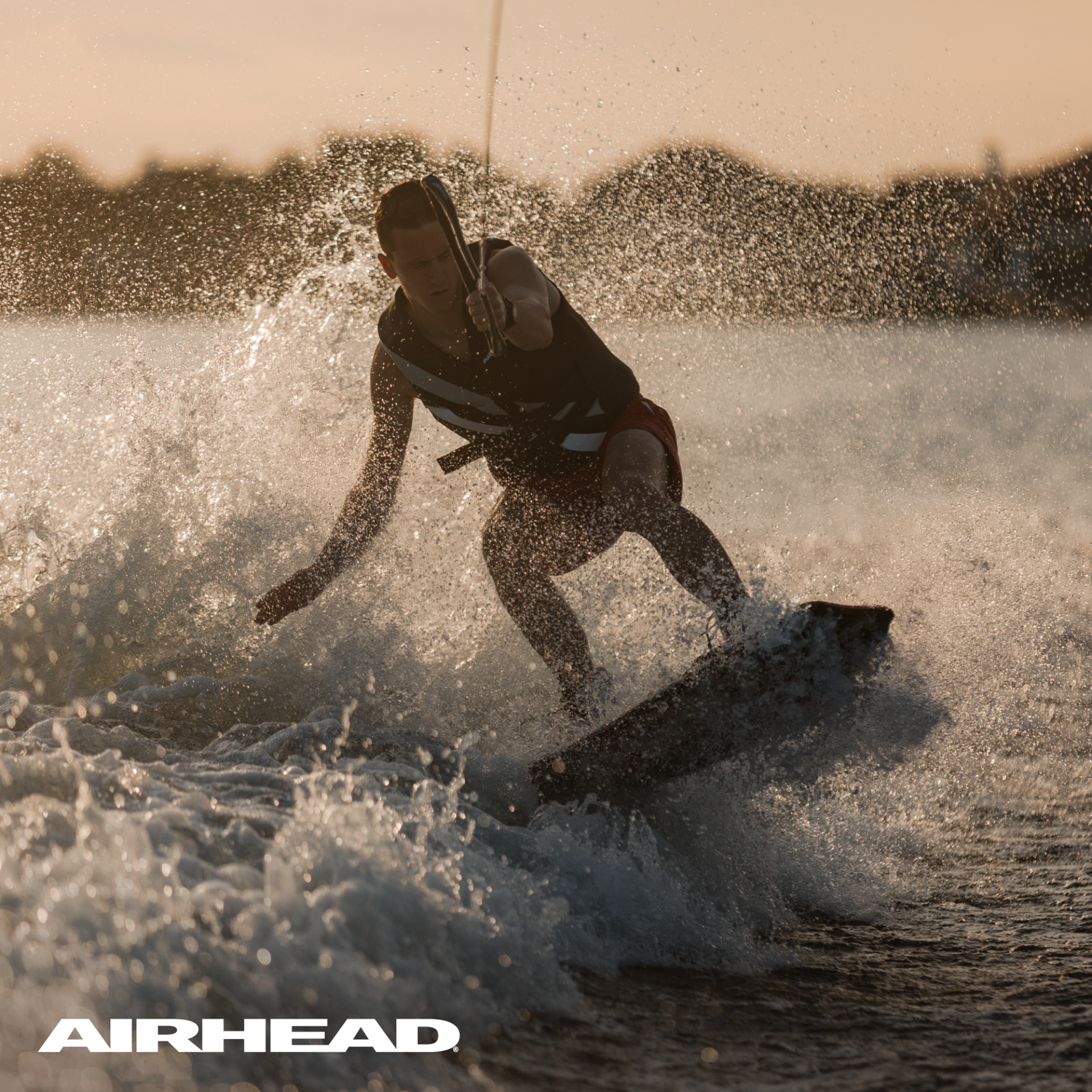 A man wakeboarding on a lake with a white Airhead logo in the bottom lefthand corner.